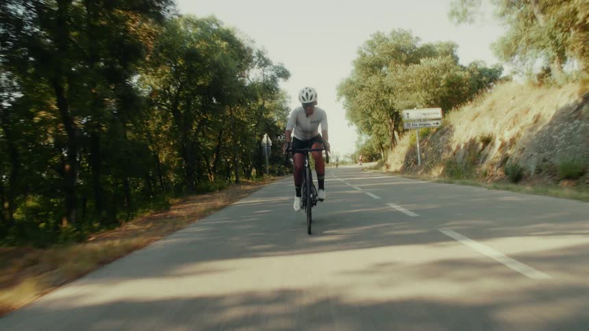 A female cyclist wearing a helmet and sunglasses rides her bike on a paved road surrounded by sun-drenched trees