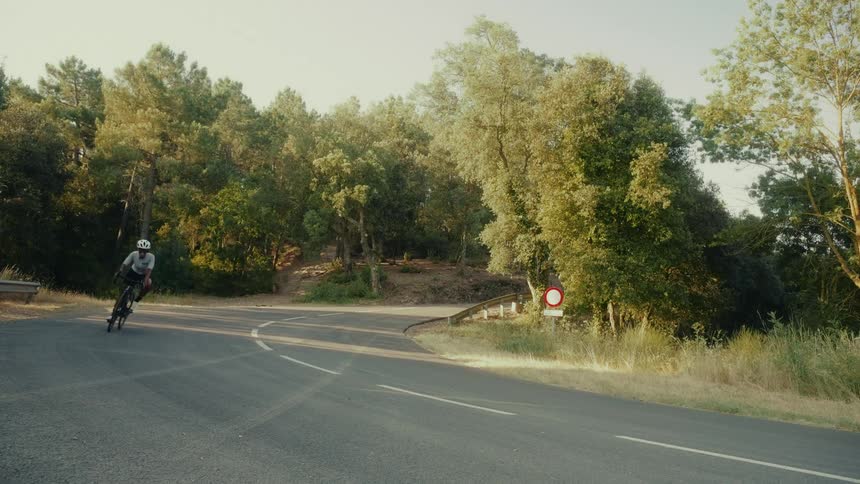 An athletic man in a helmet and professional gear rides his road bike at high speed around a curve on a country road
