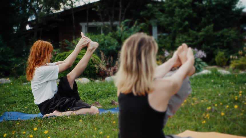 Two young women stretch in a challenging yoga pose while practicing together on a green lawn in a lush backyard
