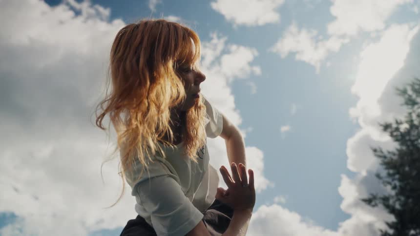 Cinemagraph of a young woman with vibrant red hair holding a contemplative pose against a bright and cloudy sky