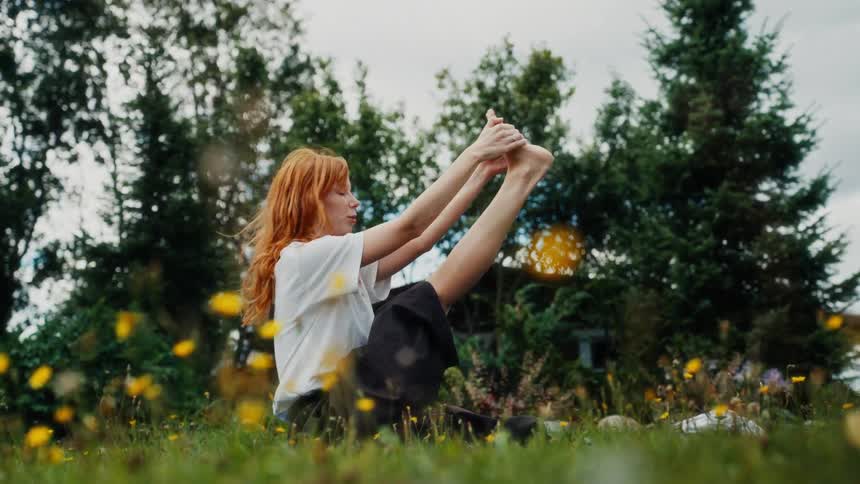 A serene young woman with long red hair holds a yoga pose while sitting on the grass in a beautiful field of yellow wildflowers