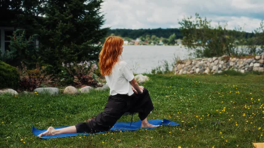 A woman with vibrant red hair performs a stretching yoga lunge pose on a blue mat in a lush green yard overlooking a serene lake