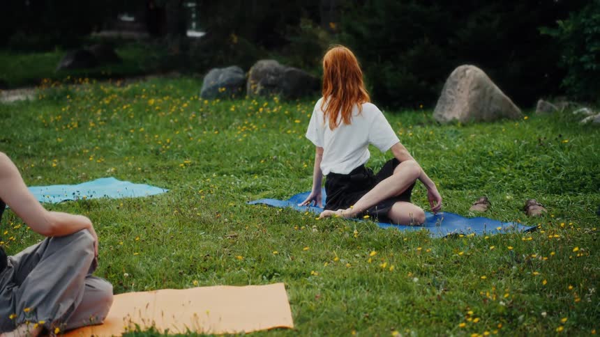 A red-haired woman holds a seated twist yoga pose during an outdoor class in a green park