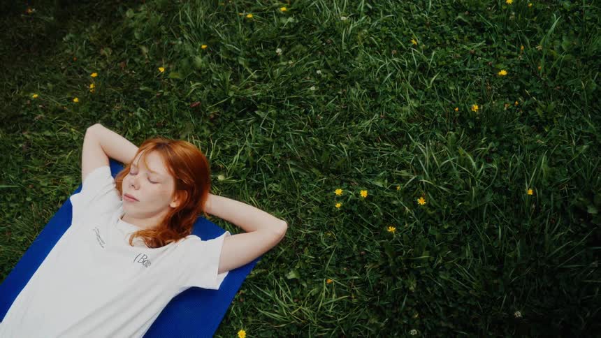A tranquil young red-haired woman with freckles peacefully rests on a blue mat in a lush green meadow with her eyes closed