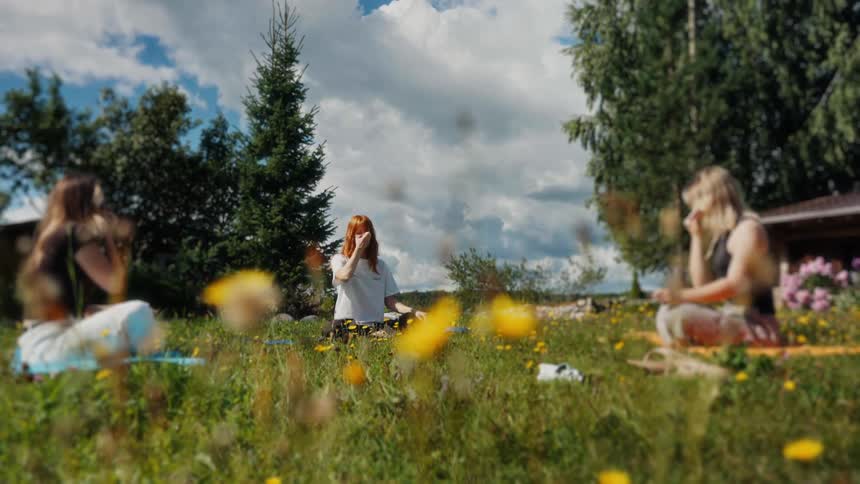 Three young women practice a breathing exercise while meditating during an outdoor yoga class in a sunny, flower-filled meadow