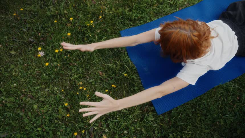Overhead view of a serene young woman with red hair performing a gentle stretching pose on a blue yoga mat in a grassy field