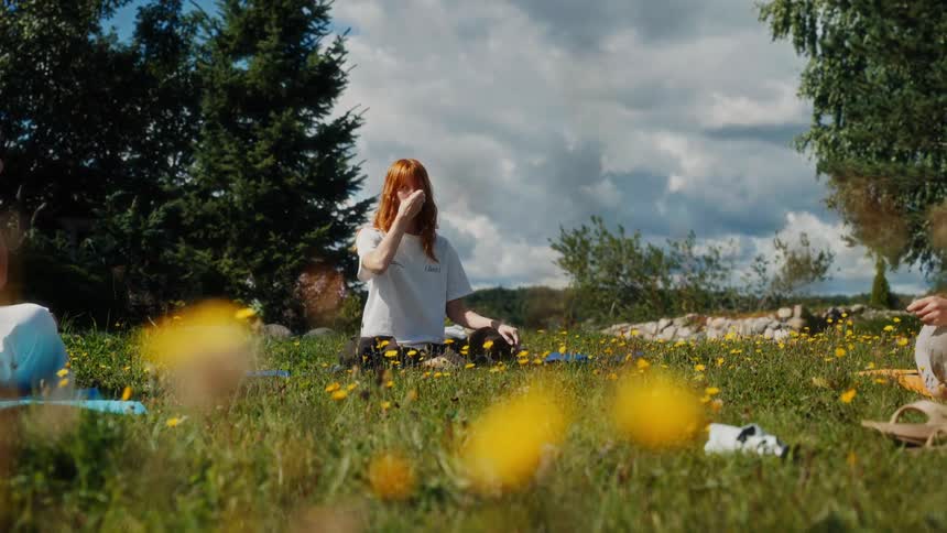 A red-haired woman practices a pranayama breathing exercise during an outdoor yoga class in a beautiful sunlit meadow