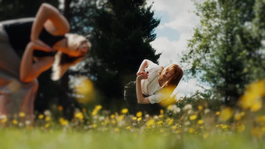 Two women perform synchronized yoga poses in slow motion in a lush green meadow filled with yellow wildflowers on a sunny day