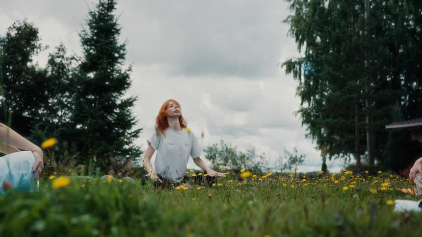 A red-haired woman leads a slow-motion yoga and meditation class in a grassy field filled with yellow dandelions
