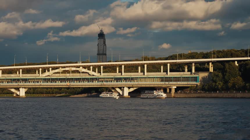 A metro train crosses the multi-level Luzhniki Bridge in Moscow with a skyscraper under construction in the background at sunset