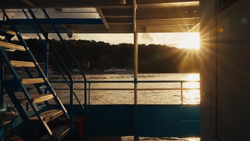 The golden sun sets over a tranquil lake, viewed from the deck of a moving passenger ferry