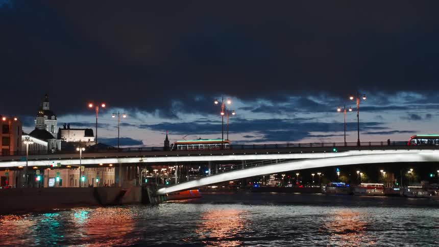 Trams cross an illuminated city bridge over a river at dusk under a dramatic cloudy sky