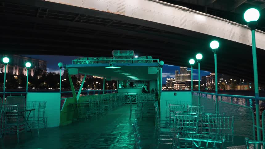 A riverboat with green lights cruises under a bridge at night with an urban cityscape in the background
