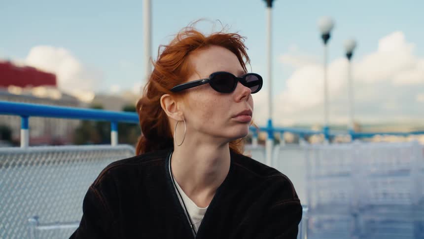 A stylish redhead woman with sunglasses sits on a boat as the wind blows through her hair in slow motion