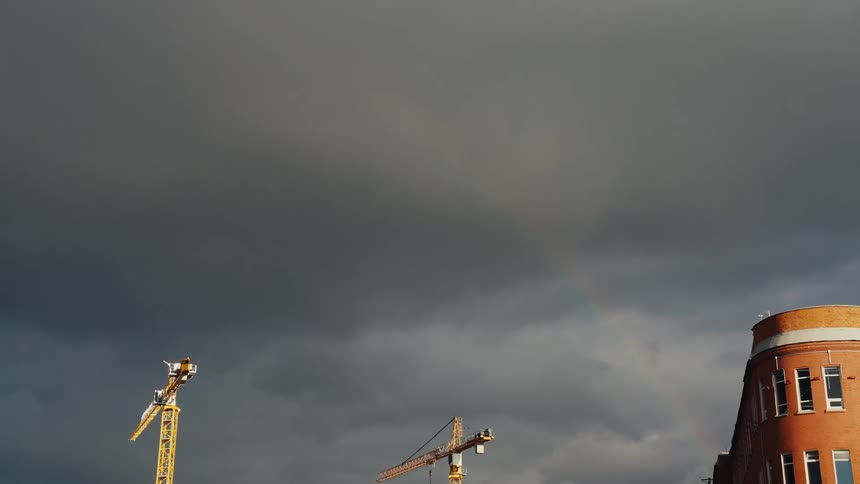 A faint rainbow arches over two yellow construction cranes against a backdrop of dark, stormy clouds in an urban setting