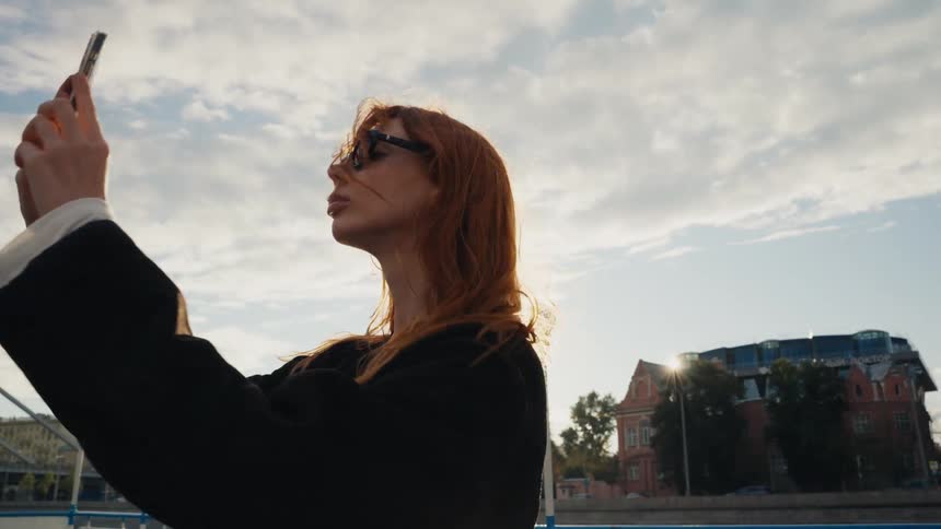 A stylish redhead in sunglasses takes photos of the city on her smartphone as the sun shines and the wind blows through her hair