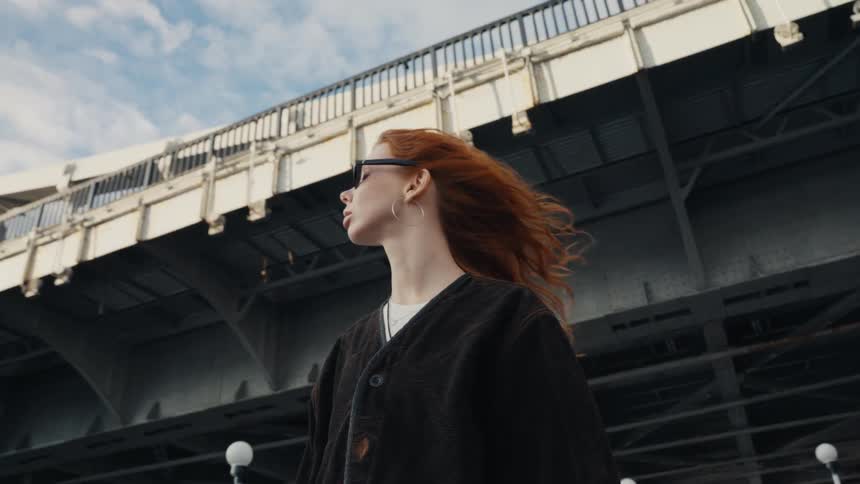 A stylish redhead in sunglasses stands beneath a large urban bridge, her hair blowing in the wind in a dramatic slow-motion shot