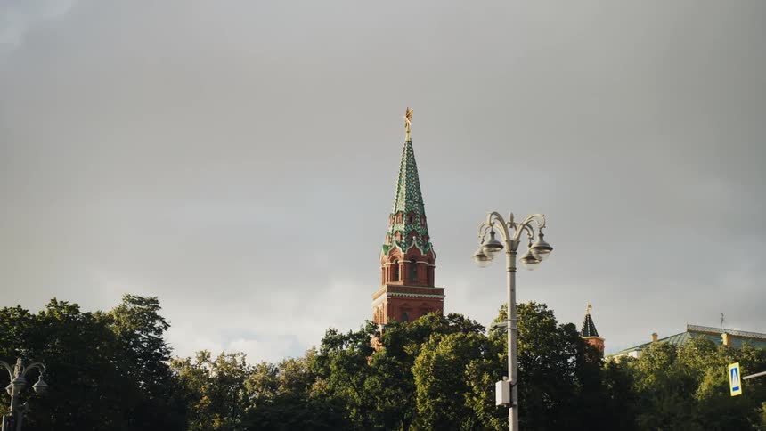 An iconic red and green spire of a Moscow Kremlin tower stands tall above the city park trees under a cloudy sky