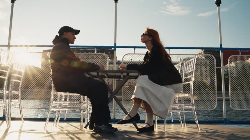 A stylish couple has a romantic conversation at a table on the deck of a boat cruising through the city at sunset