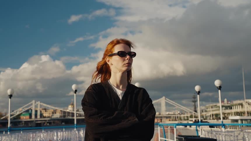 A stylish redhead woman in sunglasses stands on a moving boat, her hair blowing in the wind against a dramatic city skyline