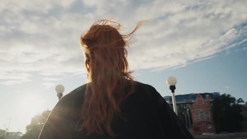 Cinematic slow motion of a woman with red hair and sunglasses walking on a city embankment as the wind blows through her hair
