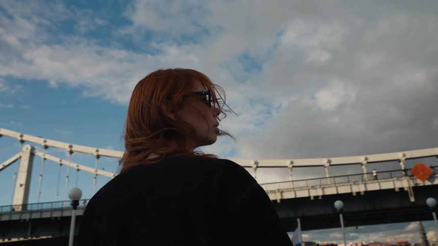 Cinematic slow motion shot of a stylish woman with red hair blowing in the wind against a city bridge and dramatic sky