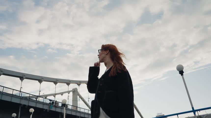 A slow-motion, low-angle shot of a contemplative red-haired woman standing near a large city bridge, with her hair blowing in the wind against a cloudy sky