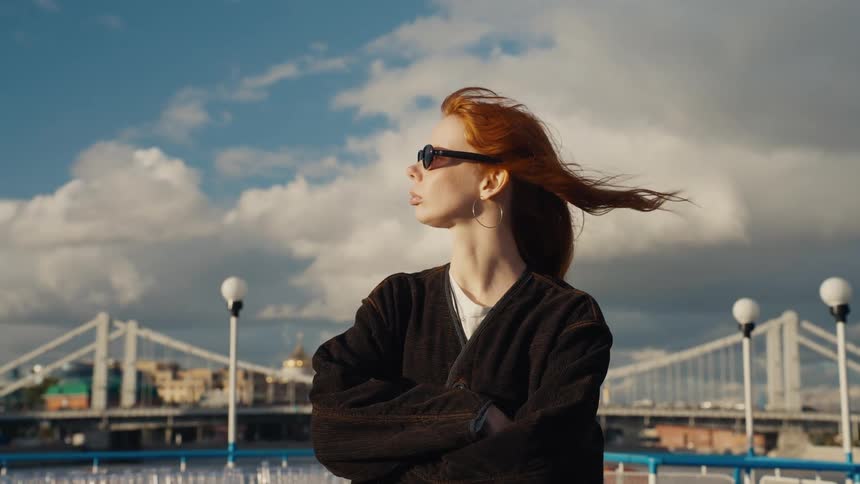 A confident woman with red hair blowing in the wind stands on a boat with a city bridge in the background