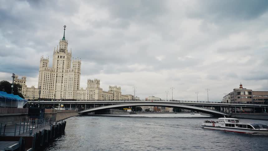 A tourist boat sails along the Moskva River past the iconic Kotelnicheskaya Embankment Building in Moscow on a cloudy day