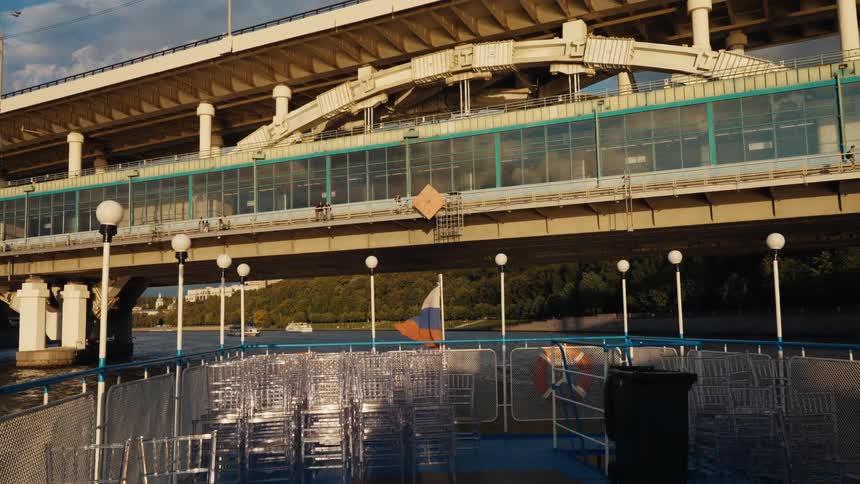 A tourist boat with a Russian flag and empty chairs sails away from the Luzhniki Metro Bridge on the Moskva River at sunset