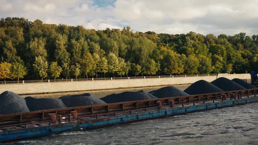 A long industrial barge loaded with piles of gravel is pushed by a tugboat along a river next to a lush, forested embankment