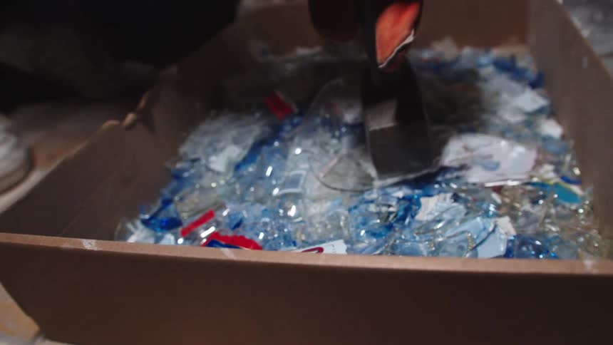 A person wearing work gloves uses a sledgehammer to smash colorful shards of glass in a cardboard box in slow motion