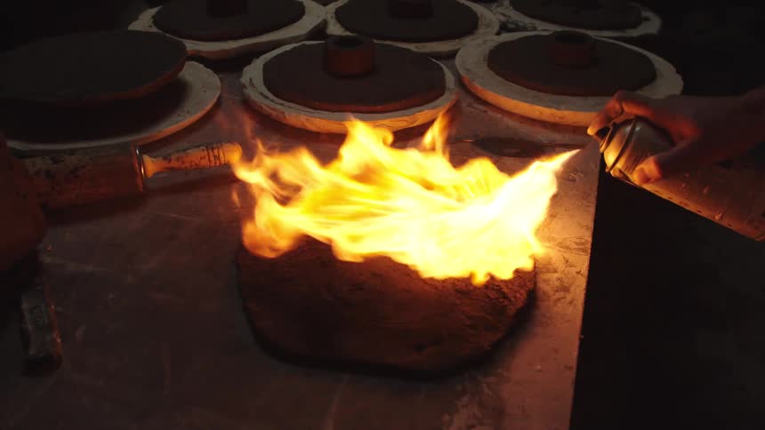An artisan carefully heats a dark clay mold with a torch in a dimly lit studio, captured in slow motion