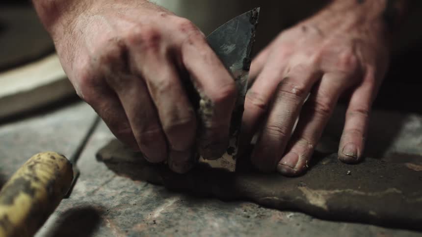 A potter carefully cuts and shapes a slab of dark clay with a metal tool in their workshop