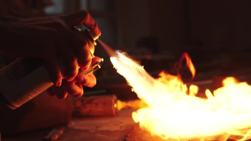 A person creates a flamethrower effect by igniting the spray from an aerosol can in a dark workshop, captured in slow motion