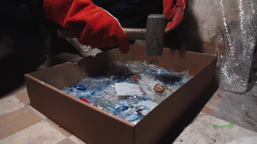 A worker in red safety gloves uses a heavy hammer to smash glass bottles into smaller pieces inside a cardboard box in slow motion
