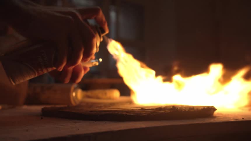 An artisan carefully fires a clay workpiece using a jet of flame from a spray can and lighter in a dark workshop, shown in slow motion