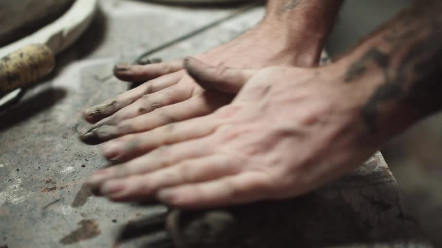 A ceramicist's tattooed hands knead, roll, and punch a lump of dark clay on a workshop table