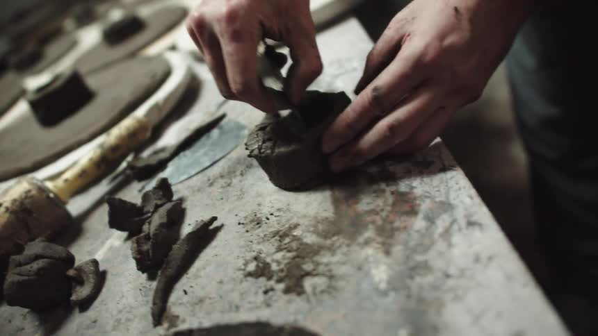 A craftsman carefully shapes and textures a small clay bowl by hand on a rustic workbench in a studio