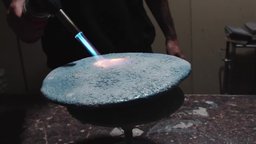 A craftsman uses a blowtorch in slow motion to treat a textured, blue surface in a workshop