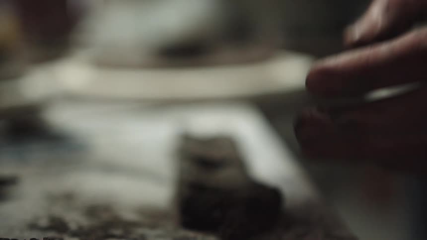A close-up slow-motion shot of an artist's hands using a sculpting tool to carefully shape a piece of dark clay in a workshop