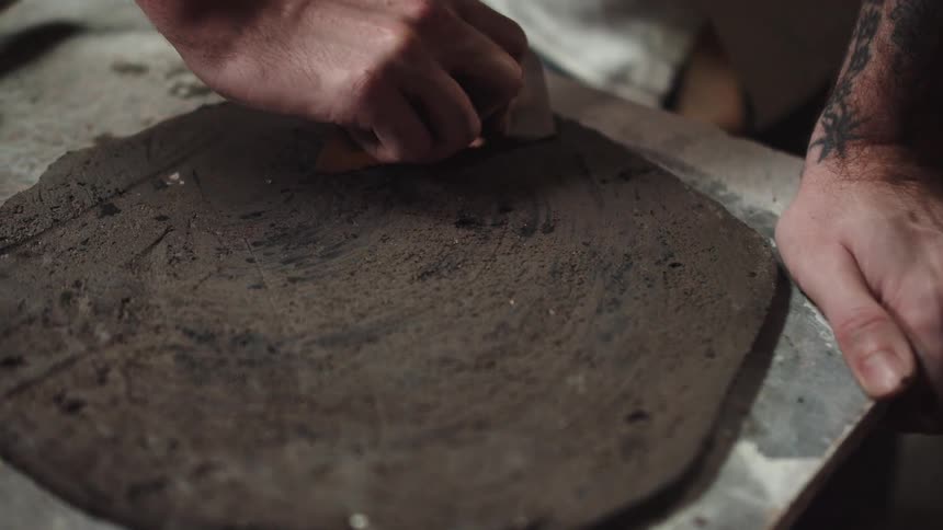 An artisan potter carefully smooths a slab of dark clay with a metal scraper tool in their workshop