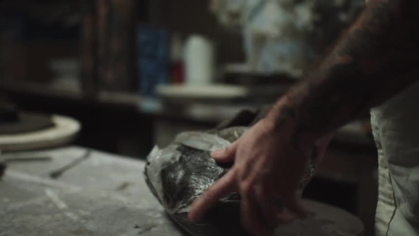 Close up of a potter's tattooed hands slowly unwrapping a large block of clay in a dimly lit studio