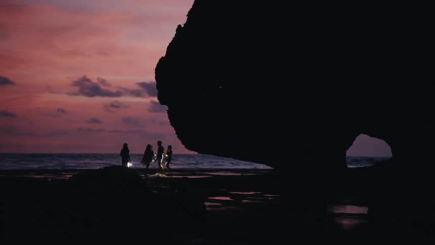 Silhouetted friends with flashlights explore a rocky beach by a large cave as the sun sets over the ocean