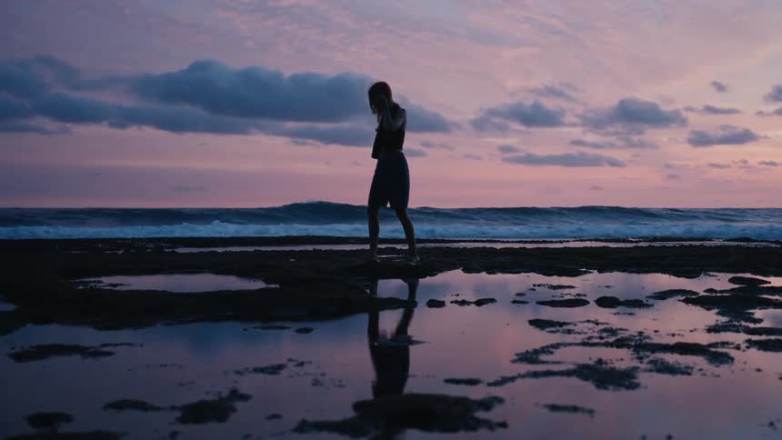 A woman walks in slow motion along a rocky shore at twilight, her silhouette reflected in tide pools