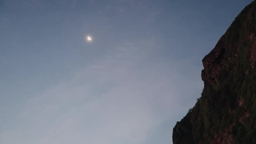 A bright crescent moon hangs in the twilight sky next to the dark silhouette of a rugged mountain cliff