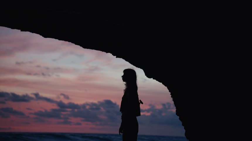 A woman's silhouette stands in a cave on the beach as her hair blows in the wind during a colorful sunset over the ocean