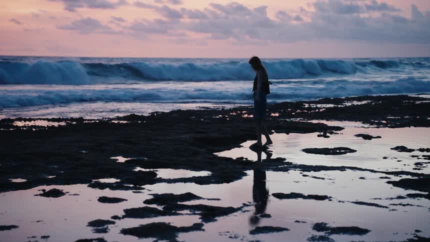 A silhouetted woman walks slowly along a rocky beach, her reflection in the tide pools under a pastel sunset sky