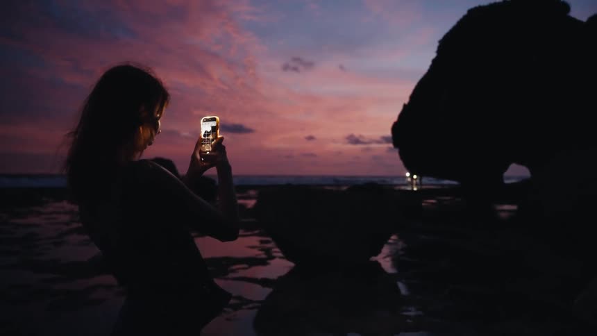 A silhouetted woman uses her smartphone to capture the stunning pink and purple sunset on a rocky beach