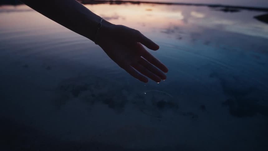 A silhouetted hand gently scoops water at dusk, letting drops fall and create ripples in slow motion